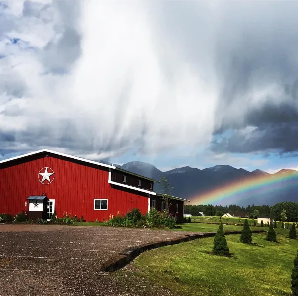 A red barn with a white star logo sits on a grassy property with mountains and a rainbow in the background.