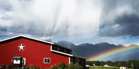 A red barn with a white star logo sits on a grassy property with mountains and a rainbow in the background.