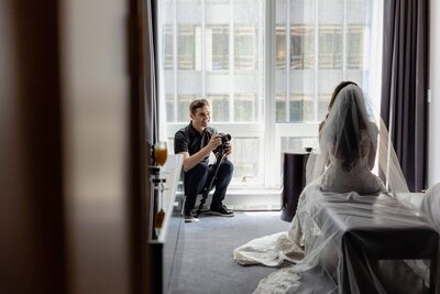 A videographer crouching by a window filming a bride in a white dress and veil inside a hotel room.