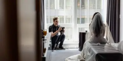 A videographer crouching by a window filming a bride in a white dress and veil inside a hotel room.