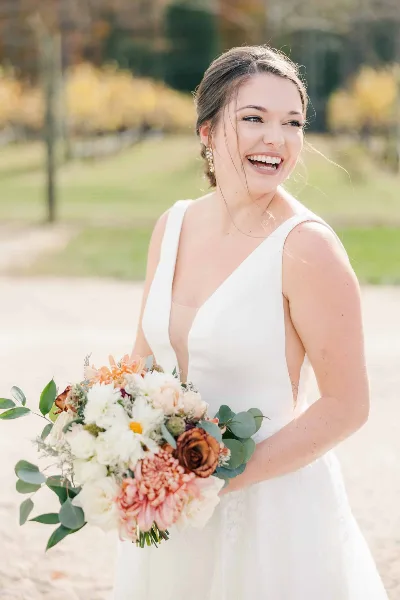 A smiling bride in a white dress holding a bouquet of flowers, showcasing her styled hair and makeup.