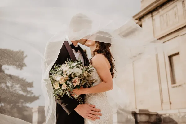 A bride and groom sharing a kiss under a veil, holding a bouquet of white and pink flowers.