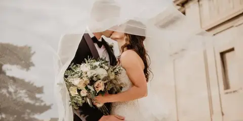 A bride and groom sharing a kiss under a veil, holding a bouquet of white and pink flowers.