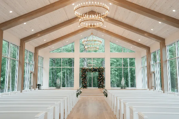 Interior view of a modern glass chapel with white pews, wooden beams, and a floral arch at the altar.