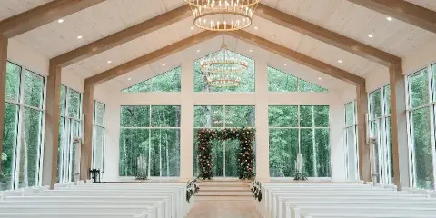 Interior view of a modern glass chapel with white pews, wooden beams, and a floral arch at the altar.