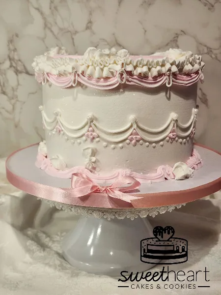 A white and pink custom cake with intricate frosting details and a ribbon bow on a white cake stand.
