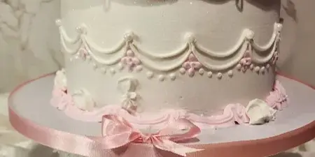 A white and pink custom cake with intricate frosting details and a ribbon bow on a white cake stand.