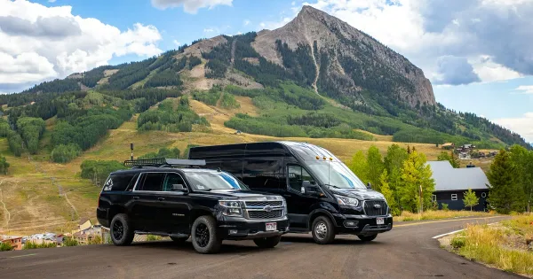 A black Chevrolet Suburban and a black Ford Transit van parked on a road with a mountain in the background.