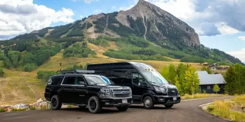 A black Chevrolet Suburban and a black Ford Transit van parked on a road with a mountain in the background.