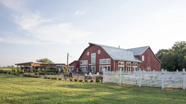 Red barn-style venue building with white picket fence, outdoor seating, and guests at an event on a grassy lawn.
