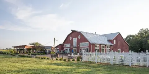Red barn-style venue building with white picket fence, outdoor seating, and guests at an event on a grassy lawn.