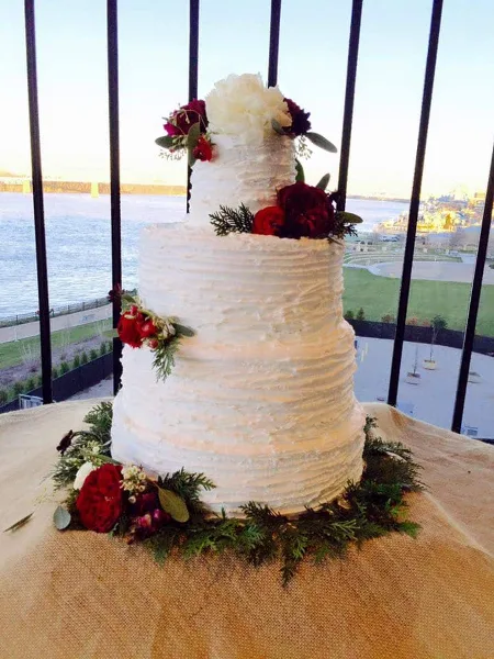 A two-tier white wedding cake decorated with red flowers and greenery, displayed on a table with a view of a river and cityscape in the background.