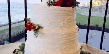 A two-tier white wedding cake decorated with red flowers and greenery, displayed on a table with a view of a river and cityscape in the background.