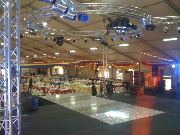 Interior of a large event tent with a stage, overhead truss lighting rig, and rows of banquet tables set for an event.