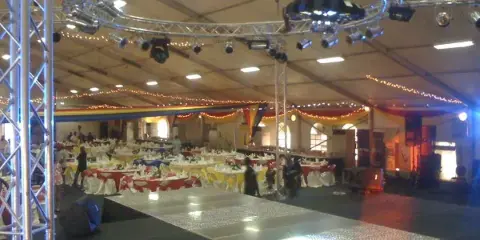 Interior of a large event tent with a stage, overhead truss lighting rig, and rows of banquet tables set for an event.