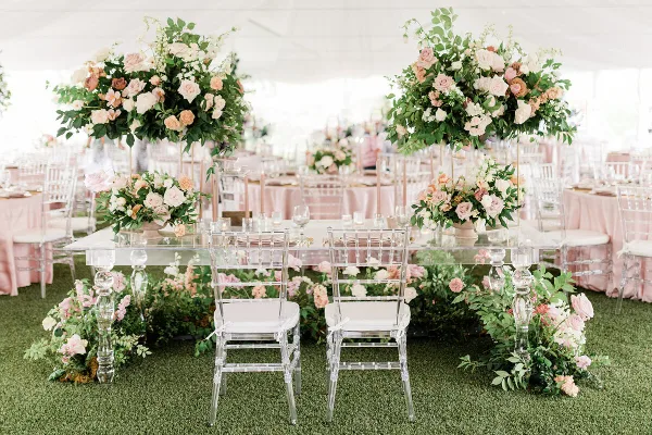 A luxury wedding reception table setting with large floral arrangements and clear chairs.