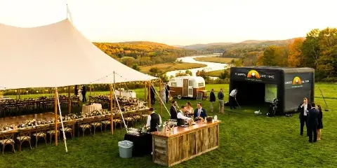 An outdoor event setup featuring a large white tent, dining tables, and a mobile golf simulator branded 'Sunrise Tee Time'.