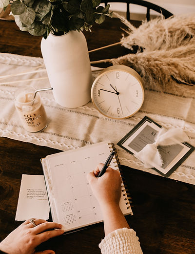 A hand writing in a planner on a table next to a clock, calendar, and candle.