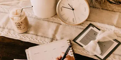 A hand writing in a planner on a table next to a clock, calendar, and candle.