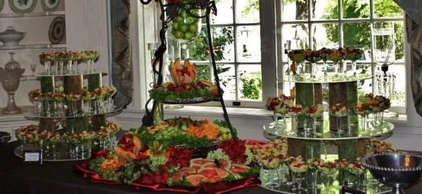 A catering buffet table featuring tiered stands of appetizers, a fruit and vegetable display, and floral arrangements in a bright room.