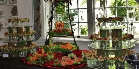 A catering buffet table featuring tiered stands of appetizers, a fruit and vegetable display, and floral arrangements in a bright room.