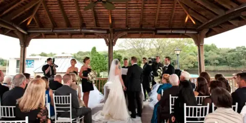 A wedding ceremony in progress at an outdoor wooden pavilion with a bride and groom standing before an officiant and seated guests.