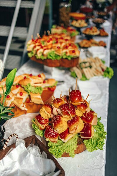 A catering buffet table displaying various appetizers including bacon-wrapped dates, sandwiches, and other prepared foods.