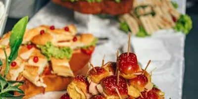 A catering buffet table displaying various appetizers including bacon-wrapped dates, sandwiches, and other prepared foods.