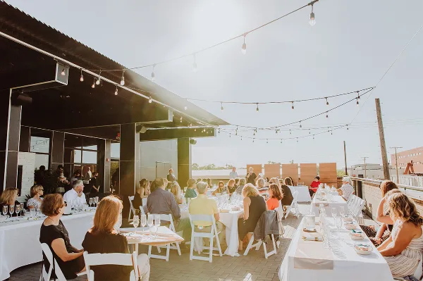 Guests seated at long white tables on an outdoor terrace with string lights overhead.