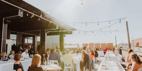 Guests seated at long white tables on an outdoor terrace with string lights overhead.