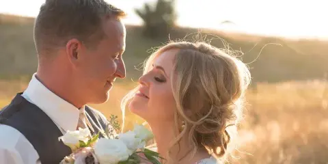A bride and groom in wedding attire sharing an intimate moment outdoors in a grassy field with golden hour lighting.