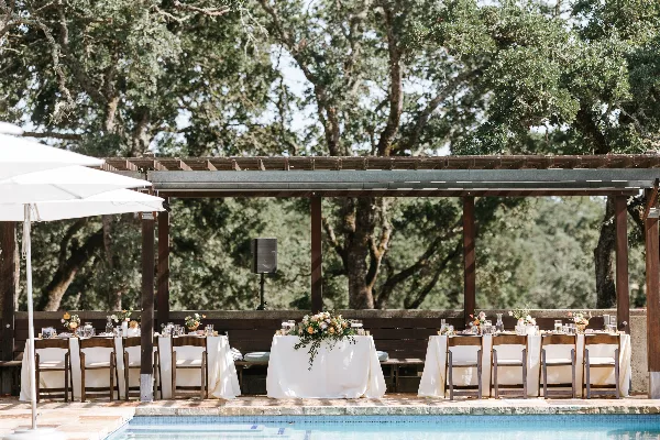 An outdoor event setup featuring wooden chairs and tables with white linens and glassware arranged under a wooden pergola.