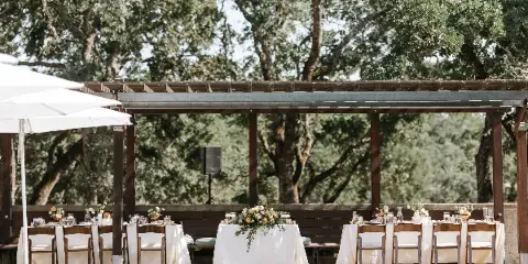 An outdoor event setup featuring wooden chairs and tables with white linens and glassware arranged under a wooden pergola.