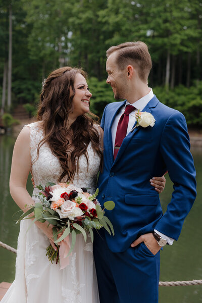 A bride and groom smiling at each other outdoors near a body of water, with the bride holding a bouquet.