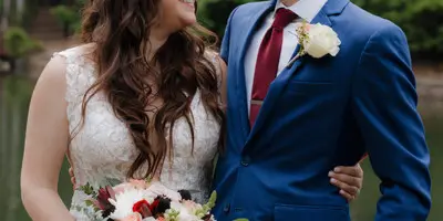 A bride and groom smiling at each other outdoors near a body of water, with the bride holding a bouquet.
