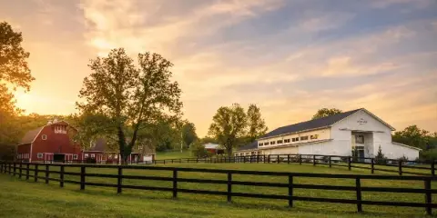 A wide shot of a farm at sunset featuring a red barn on the left and a white event venue building labeled 'The Artisan' on the right, separated by a grassy field and black fencing.