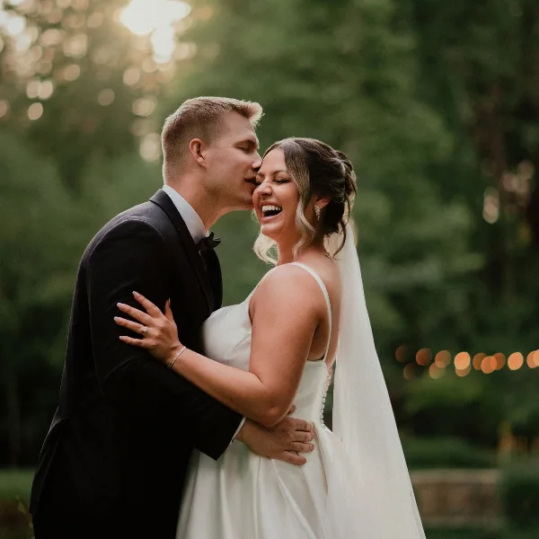 A bride with soft glam makeup and an updo hairstyle shares a joyful moment with her groom outdoors.