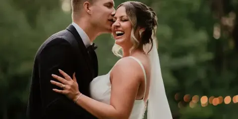 A bride with soft glam makeup and an updo hairstyle shares a joyful moment with her groom outdoors.