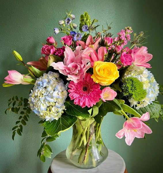 A vibrant floral arrangement featuring pink lilies, roses, and hydrangeas in a clear glass vase against a green background.