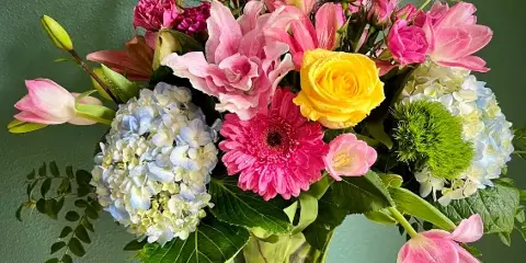 A vibrant floral arrangement featuring pink lilies, roses, and hydrangeas in a clear glass vase against a green background.