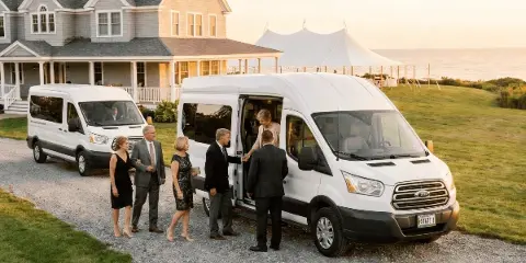 A white Ford Transit van with a driver assisting formally dressed guests at a coastal wedding venue with a white tent in the background.