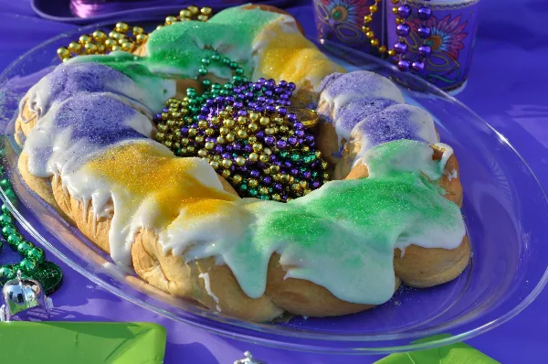 A traditional King Cake with white icing and purple, green, and gold sugar on a glass plate.