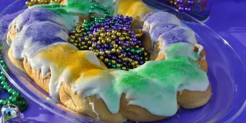 A traditional King Cake with white icing and purple, green, and gold sugar on a glass plate.