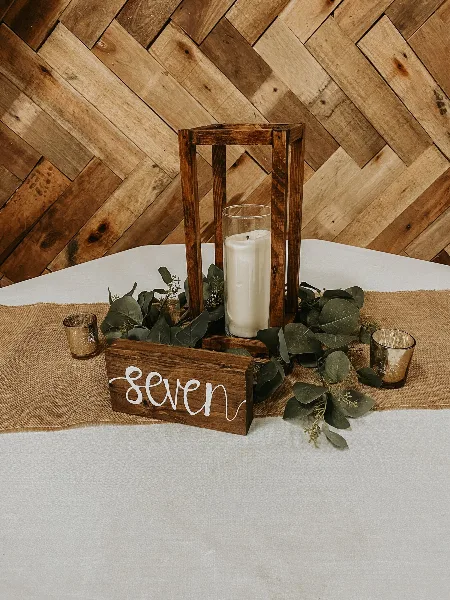 A rustic wedding table setting with a wooden lantern, white pillar candle, eucalyptus greenery, and a wooden sign reading 'seven' on a burlap runner.