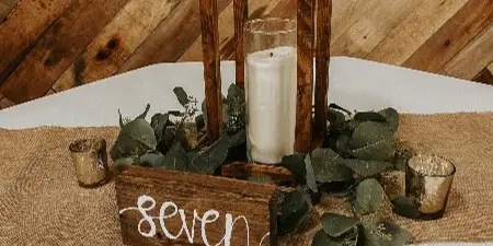 A rustic wedding table setting with a wooden lantern, white pillar candle, eucalyptus greenery, and a wooden sign reading 'seven' on a burlap runner.