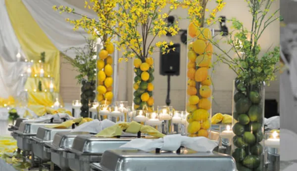 A catering buffet line with stainless steel chafing dishes, yellow floral centerpieces, and candles at a wedding event.
