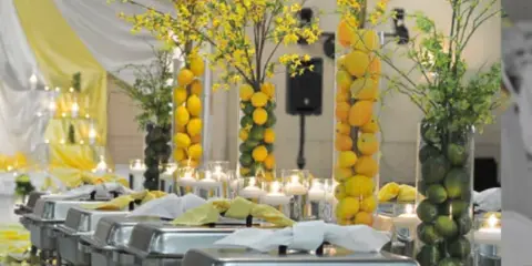 A catering buffet line with stainless steel chafing dishes, yellow floral centerpieces, and candles at a wedding event.