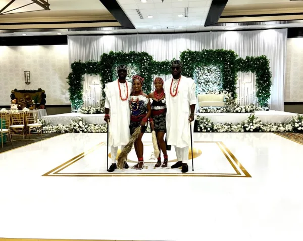 A wedding couple and family posing in front of a decorated stage with white draping, floral arches, and a dance floor.