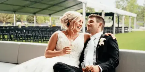 A bride and groom sitting together at a wedding reception, smiling and holding drinks.