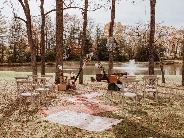 Outdoor wedding ceremony setup with a floral arch, rustic chairs, and rugs on a lawn near a pond.